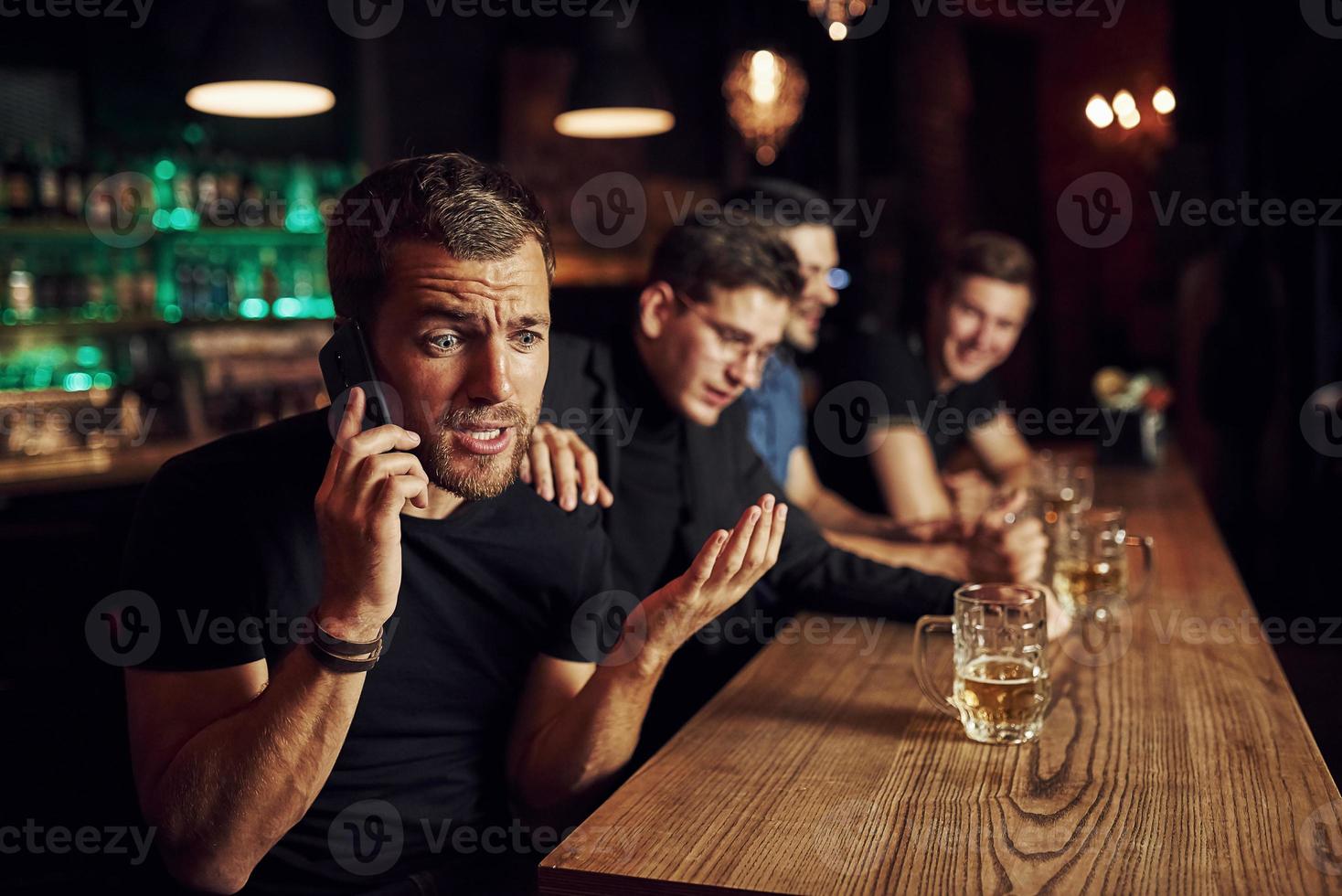 el hombre habla por teléfono. tres aficionados al deporte en un bar viendo fútbol. con cerveza en las manos foto