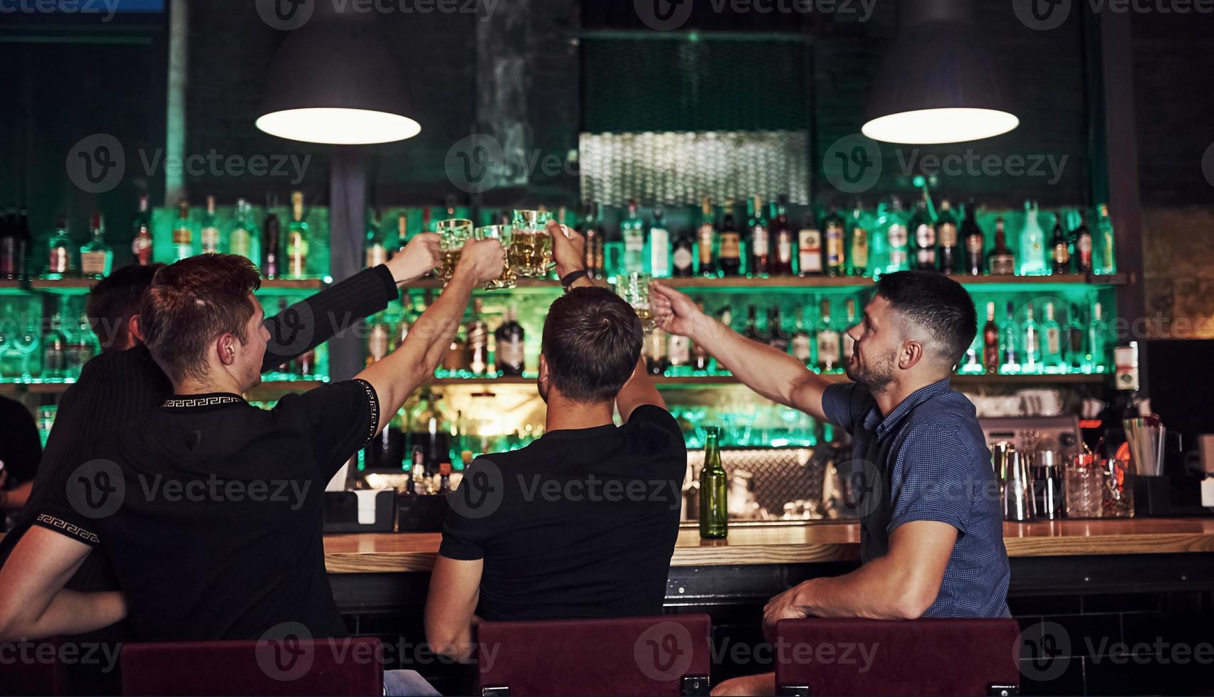 Many of bottles with alcohol on the shelves. Friends resting in the pub with beer in hands. Having conversation photo