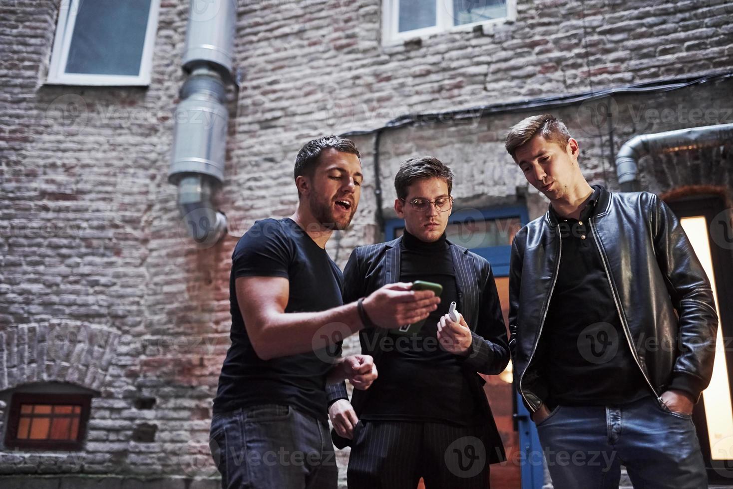 With smartphone. Some funny stuff. Three male friends stands outside the bar. Having conversation. In nice clothes photo