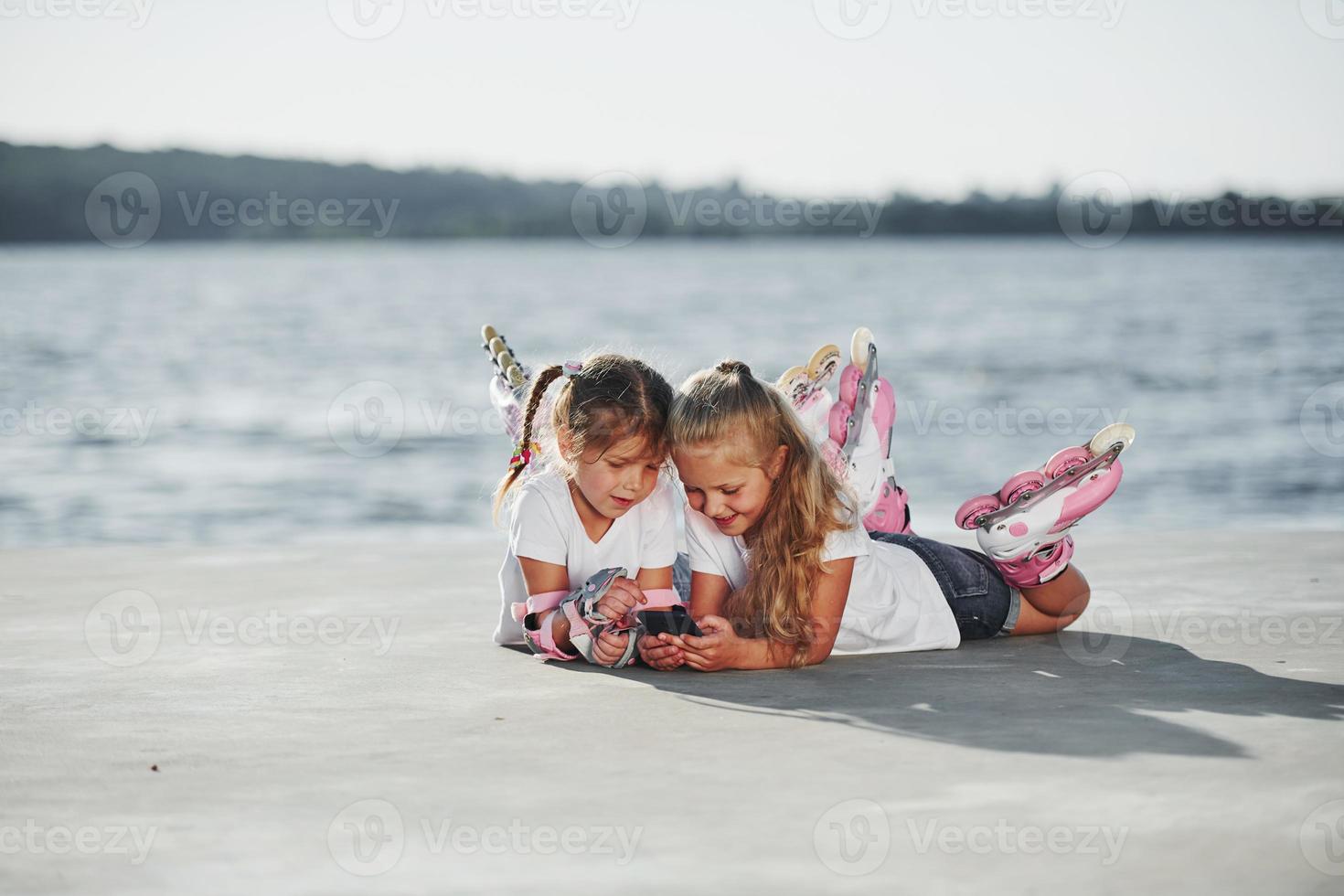 usando un teléfono inteligente. dos niñas pequeñas con patines al aire libre cerca del lago al fondo foto