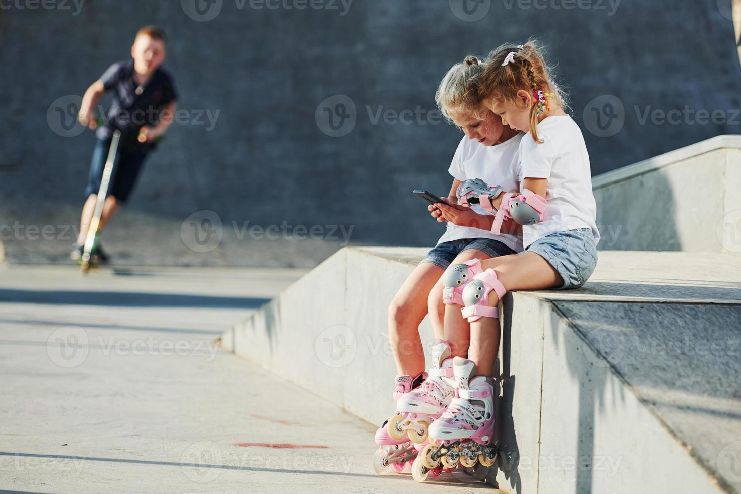 Using smartphone. On the ramp for extreme sports. Two little girls with roller skates outdoors have fun photo