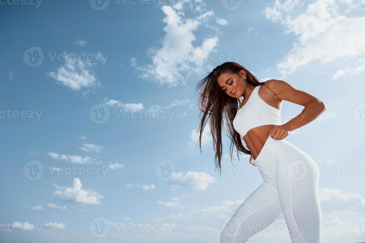 Brunette fitness woman stands outdoors in white sportive clothes against cloudy sky at daytime photo