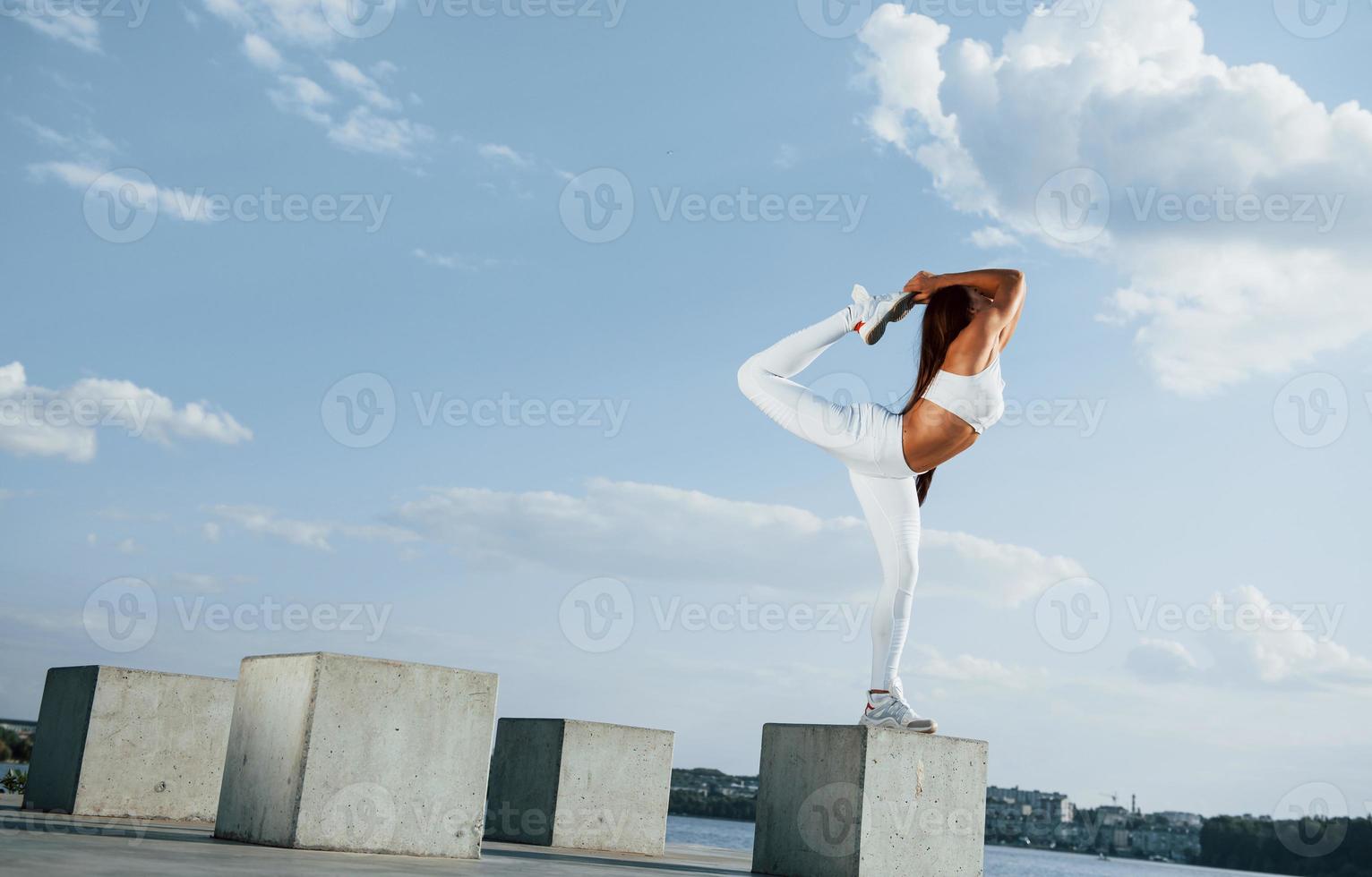 On the cement cube. Shot of sportive woman doing fitness exercises near the lake at daytime photo