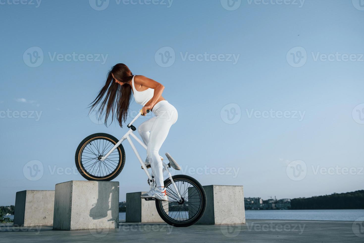 Photo in motion. Doing stunts. Female rider is on the bike at daytime near the lake. Fitness woman in sportive clothes