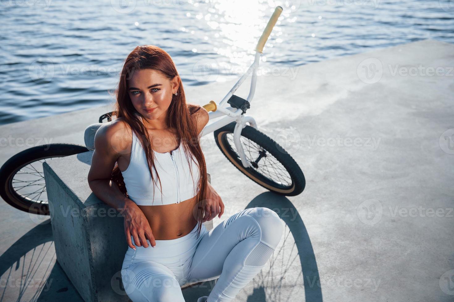 Near the bike. Fitness woman having a rest near the lake at daytime. Beautiful sunlight photo