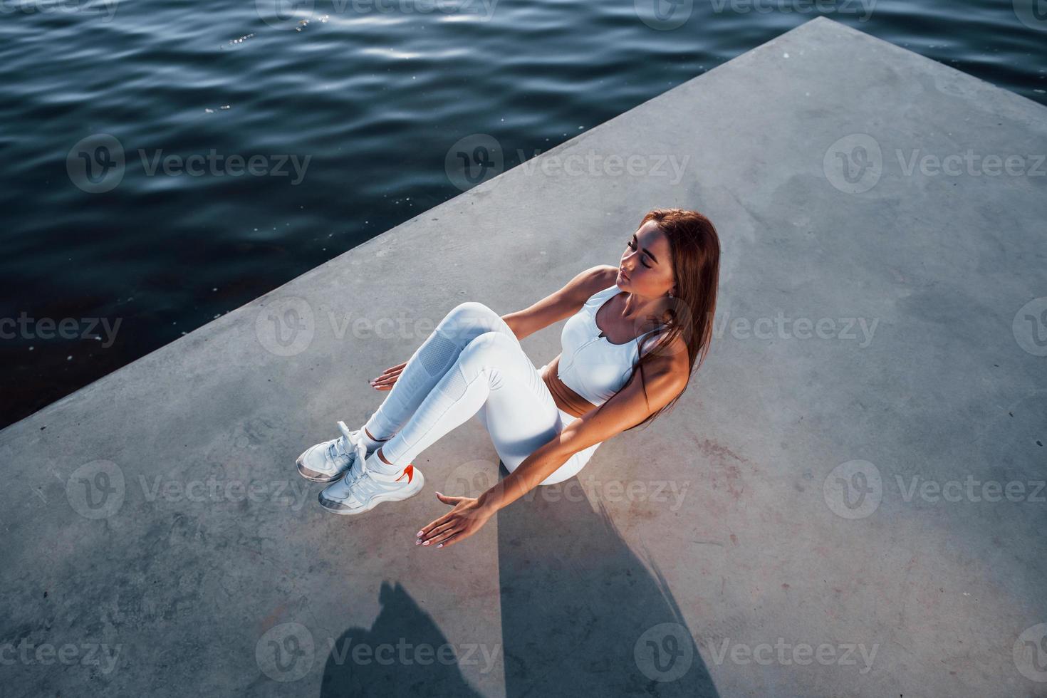 Doing abs. Top view. Shot of sportive woman doing fitness exercises near the lake at daytime photo