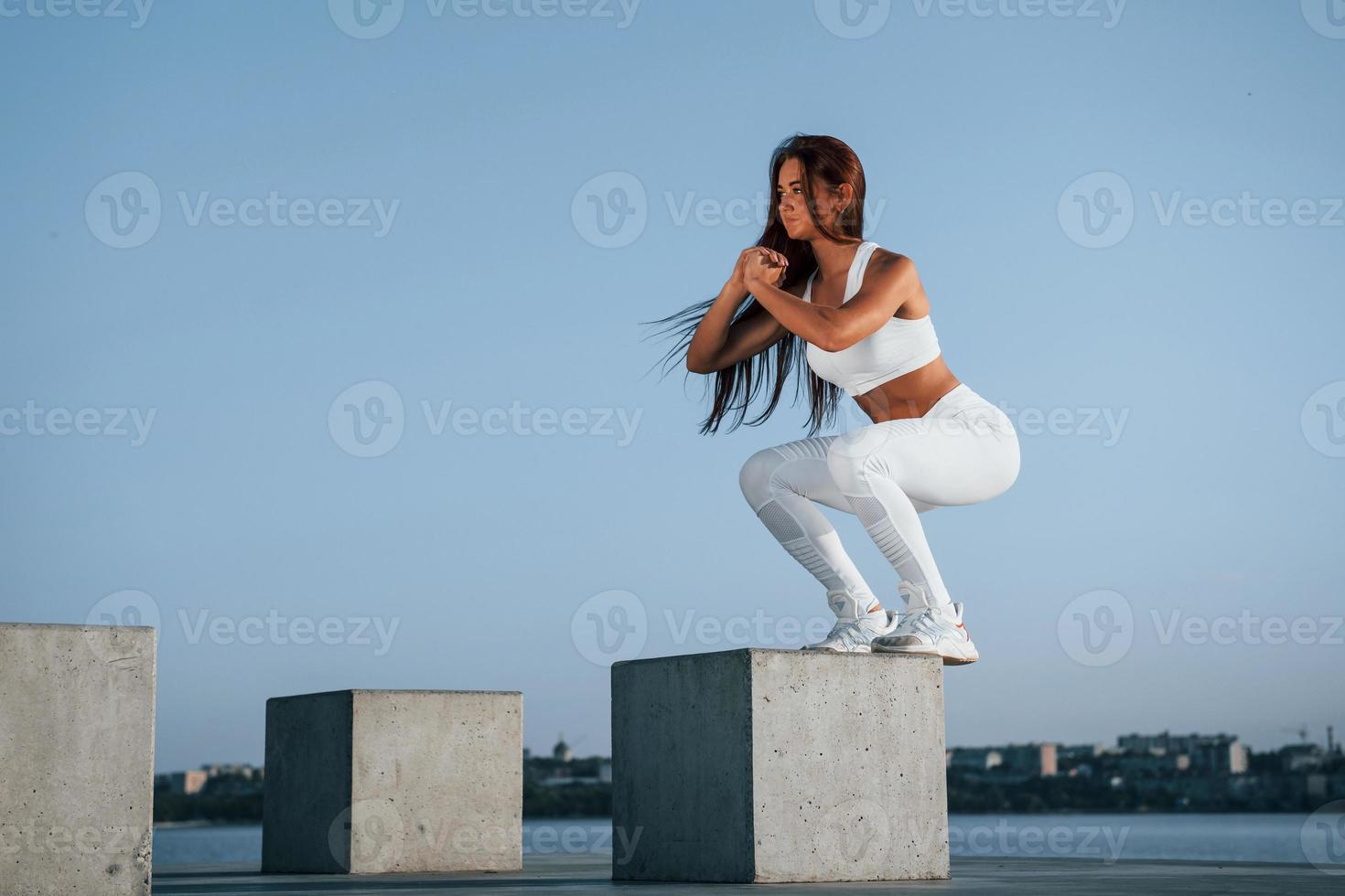 Doing squats on the cement cube. Shot of sportive woman doing fitness exercises near the lake at daytime photo