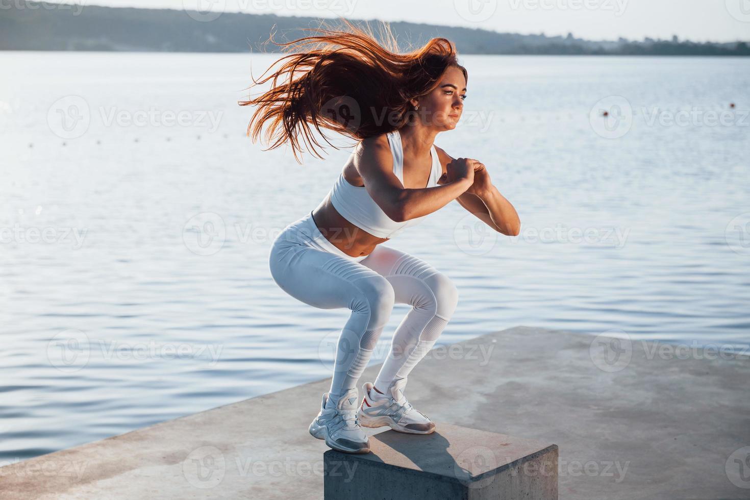 Doing squats on the cement cube. Shot of sportive woman doing fitness exercises near the lake at daytime photo