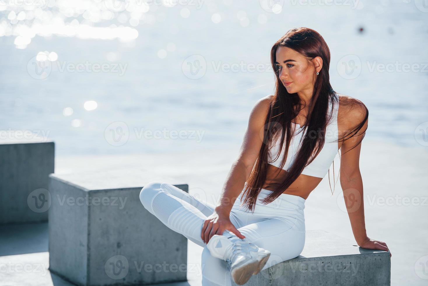 Sits on cement cube. Fitness woman having a rest near the lake at daytime. Beautiful sunlight photo