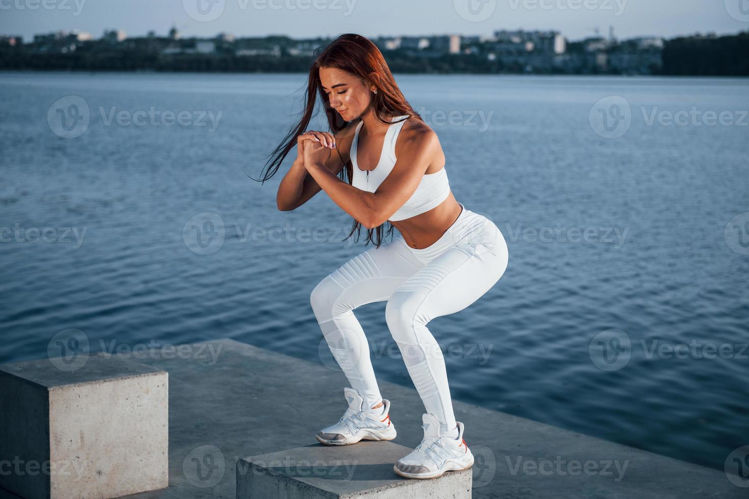 Doing squats on the cement cube. Shot of sportive woman doing fitness exercises near the lake at daytime photo