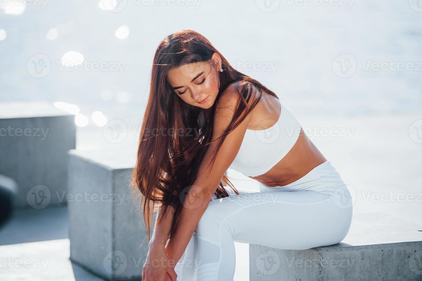 Sits on cement cube. Fitness woman having a rest near the lake at daytime. Beautiful sunlight photo