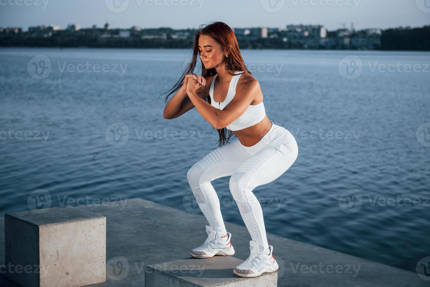 Doing squats on the cement cube. Shot of sportive woman doing fitness exercises near the lake at daytime photo