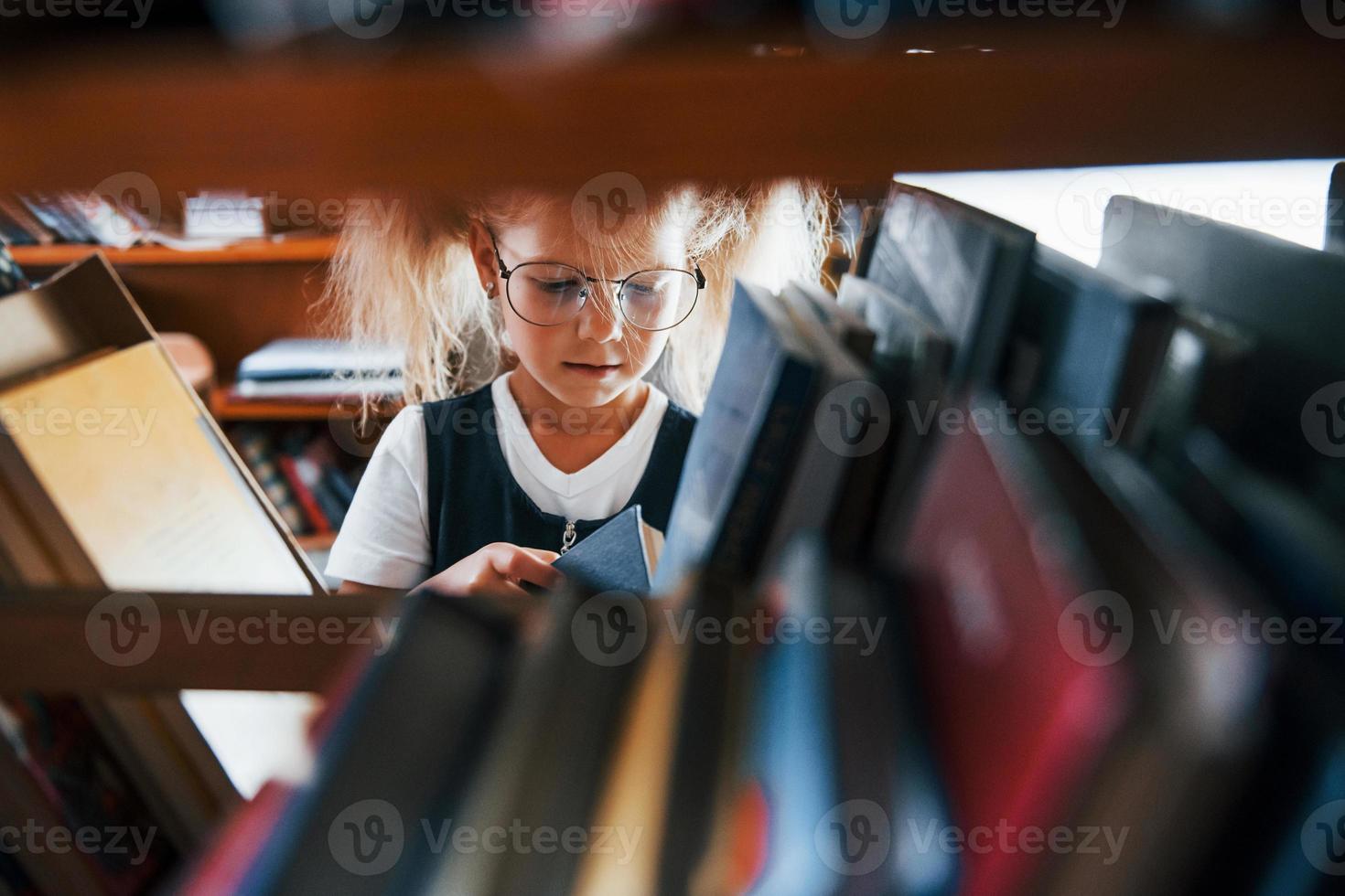 Little girl in glasses searching for a book in the library. Conception of education photo