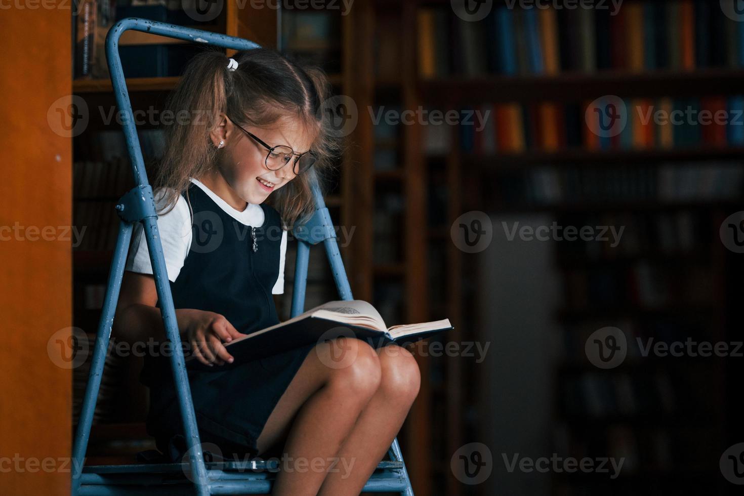 concentración en la lectura. niña de la escuela en la escalera en la biblioteca llena de libros. concepción de la educación foto