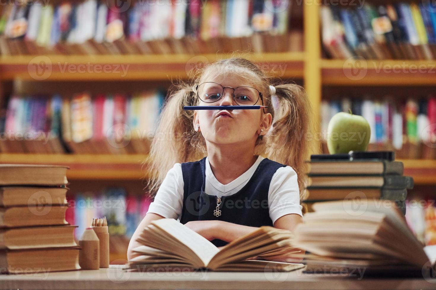 lápiz en los labios. linda niña con coletas está en la biblioteca. manzana en los libros foto