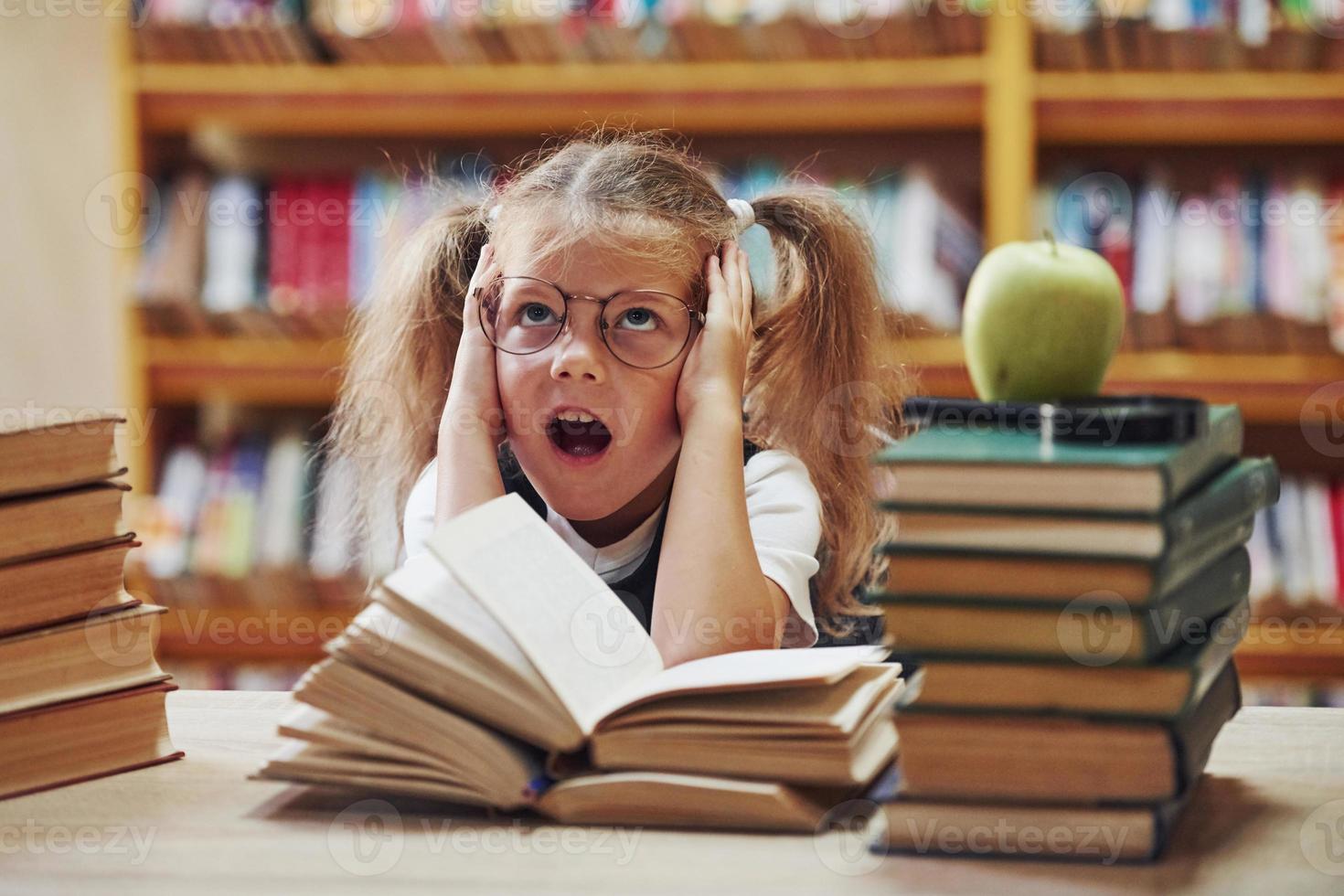 Feels excited. Cute little girl with pigtails is in the library. Apple on the books photo