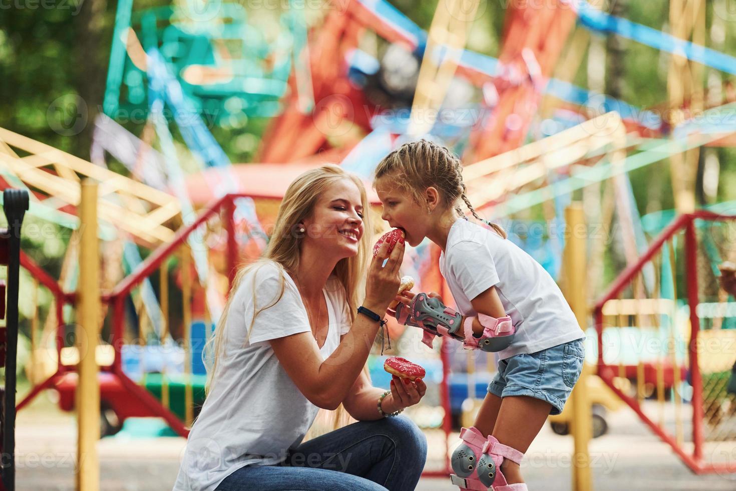 comiendo donas una niña alegre en patines y su madre se divierten juntos en el parque cerca de las atracciones foto