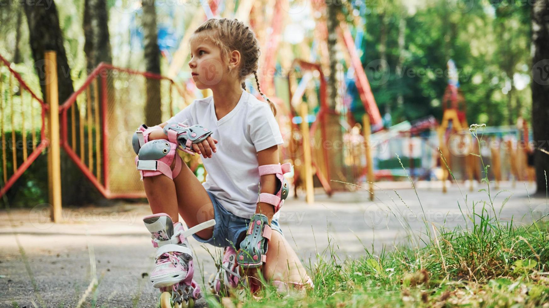 Sits on the green grass. Cheerful little girl on roller skates have a good time in the park near attractions photo