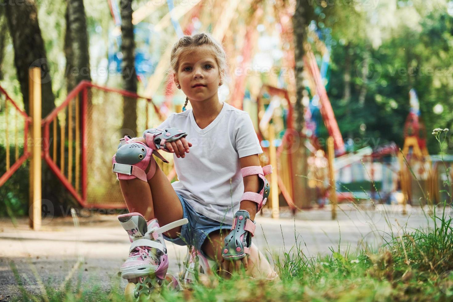 Sits on the green grass. Cheerful little girl on roller skates have a good time in the park near attractions photo