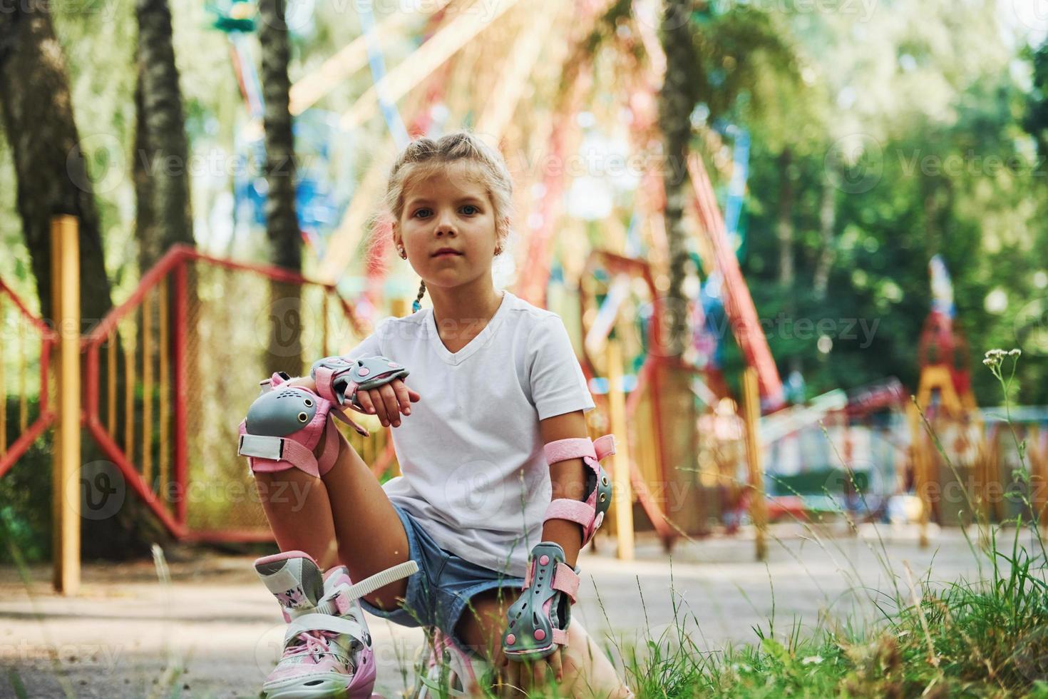 Sits on the green grass. Cheerful little girl on roller skates have a good time in the park near attractions photo