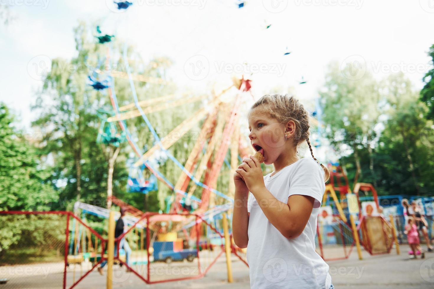 Cute little girl eats ice cream in the park at daytime near attractions photo