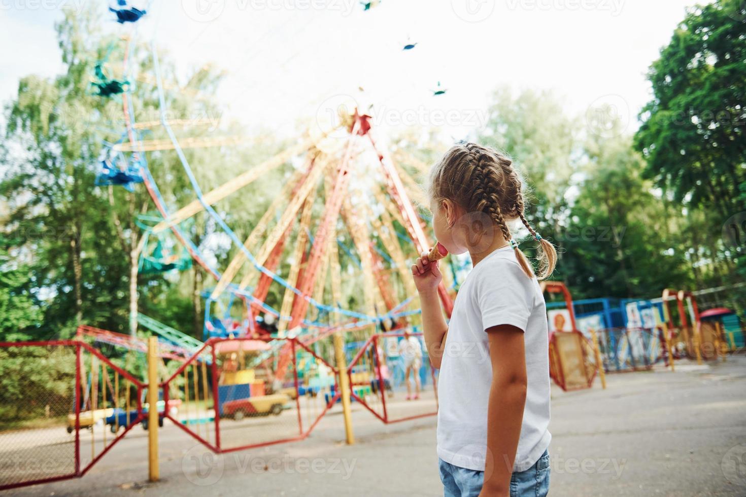 Cute little girl eats ice cream in the park at daytime near attractions photo