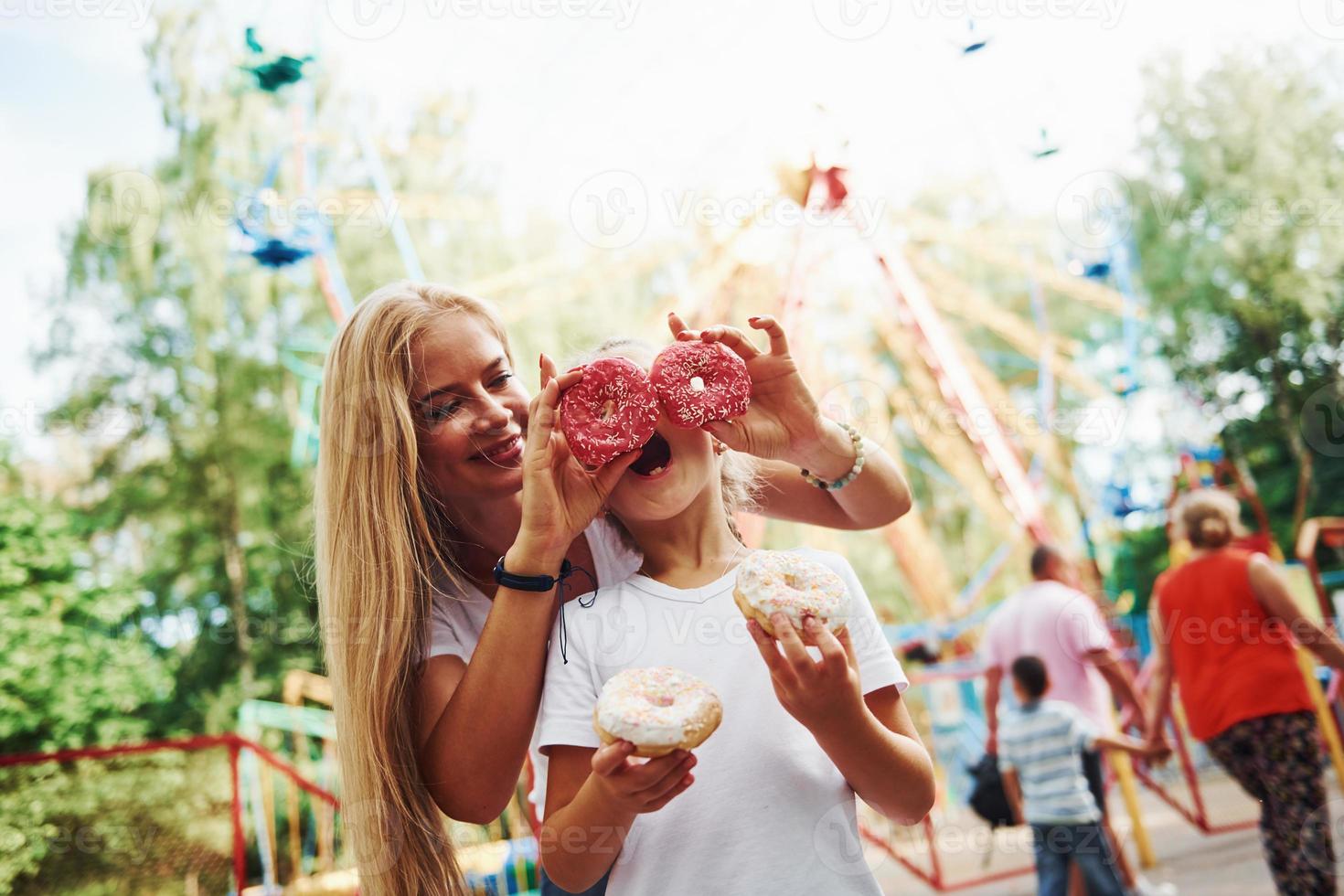 Having fun with donuts. Cheerful little girl her mother have a good time in the park together near attractions photo