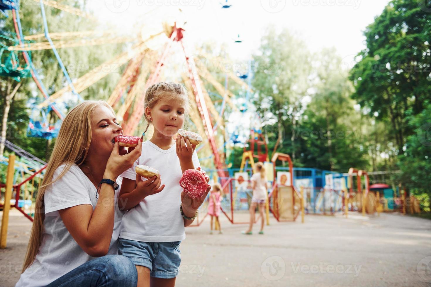 comiendo donas una niña alegre en patines y su madre se divierten juntos en el parque cerca de las atracciones foto