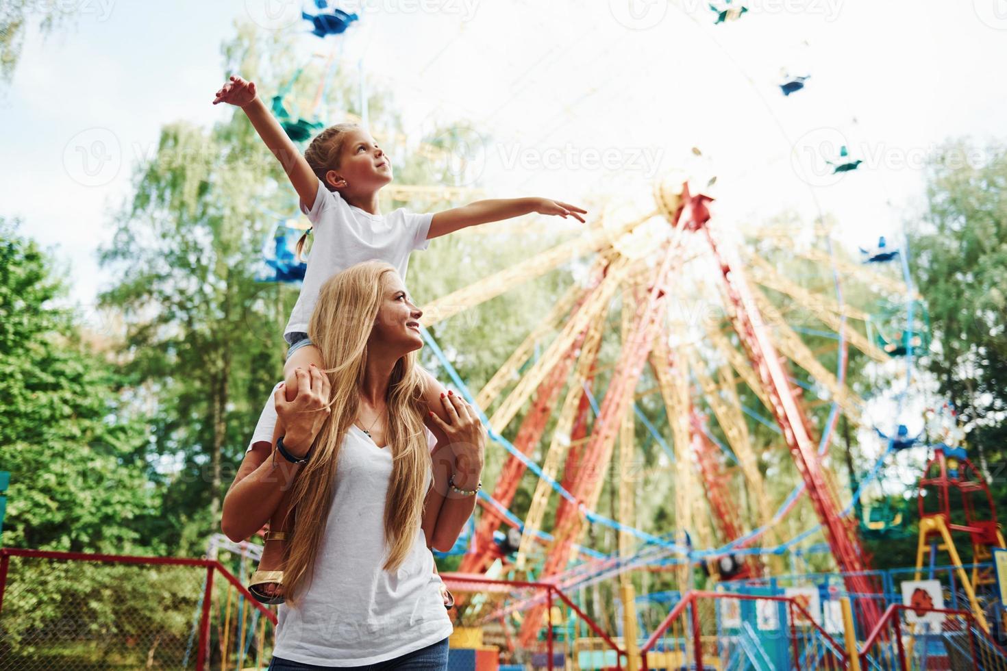 Daughter sits on the shoulders. Cheerful little girl her mother have a good time in the park together near attractions photo