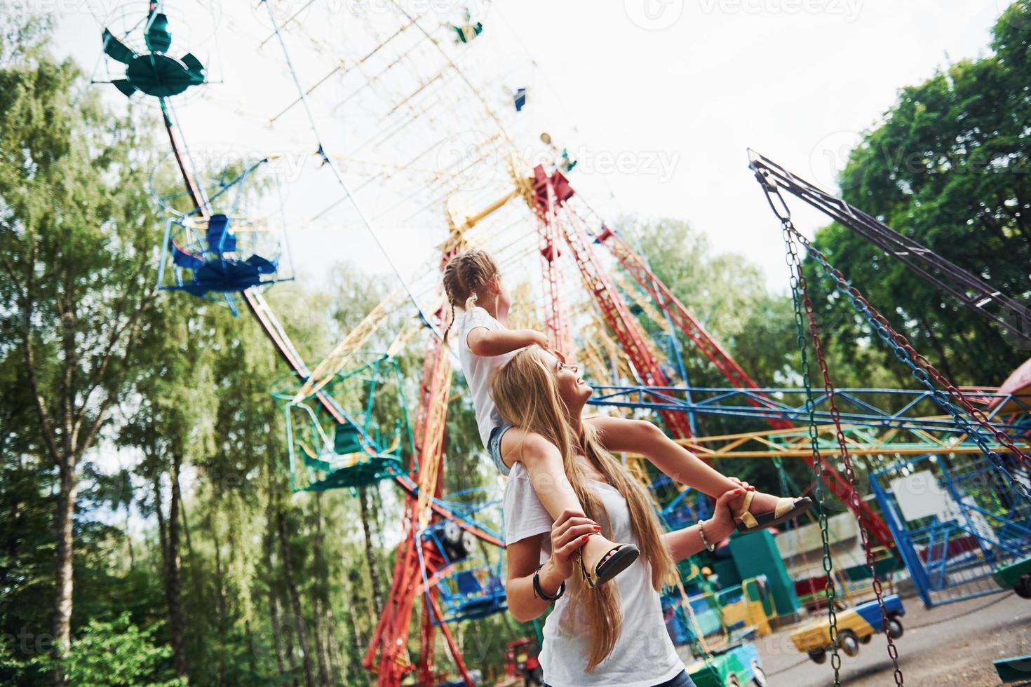 caminando con la hija en los hombros. niña alegre su madre se divierten juntos en el parque cerca de las atracciones foto