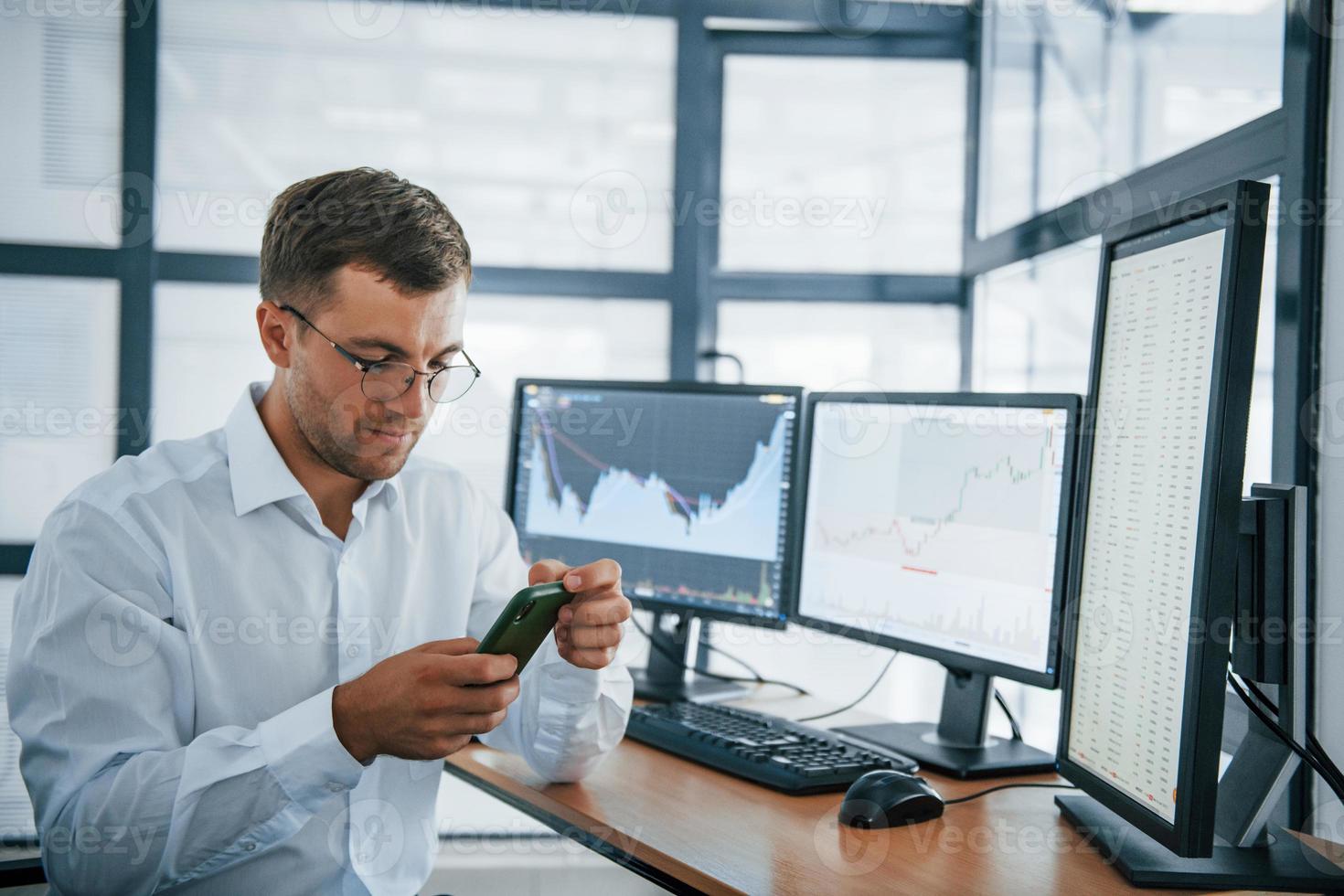 Using smartphone. Young businessman in formal clothes is in office with multiple screens. Conception of exchange and money photo