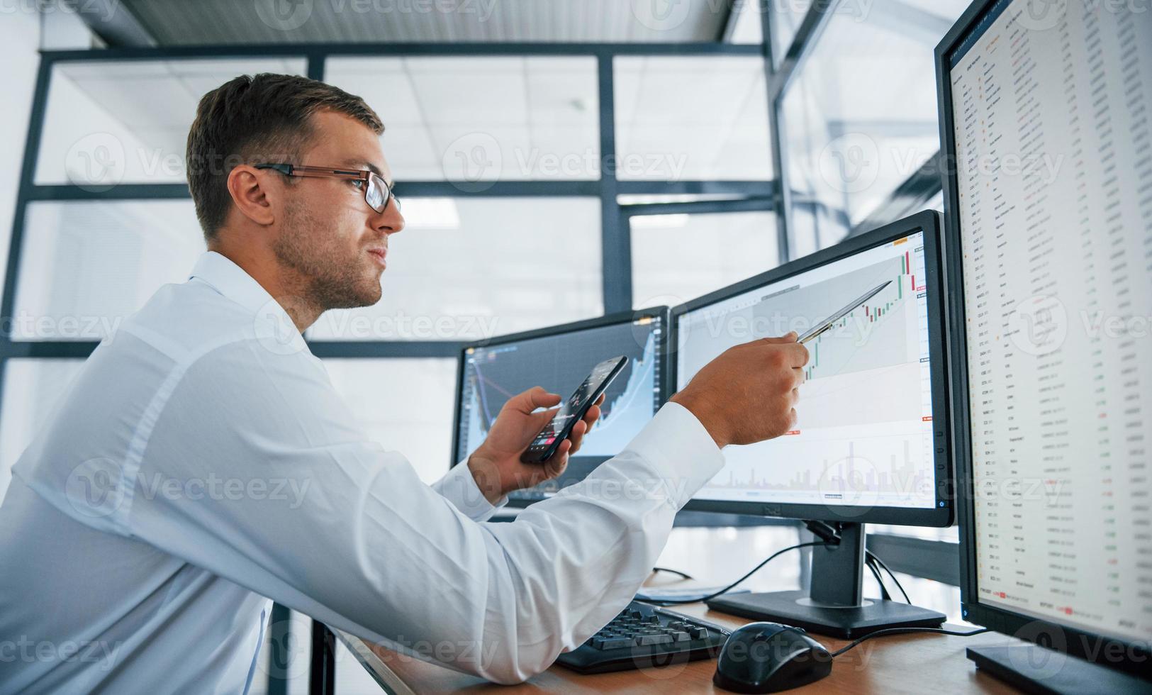 With phone and pen. Analysing graphs. Young businessman in formal clothes is in office with multiple screens. Conception of exchange and money photo