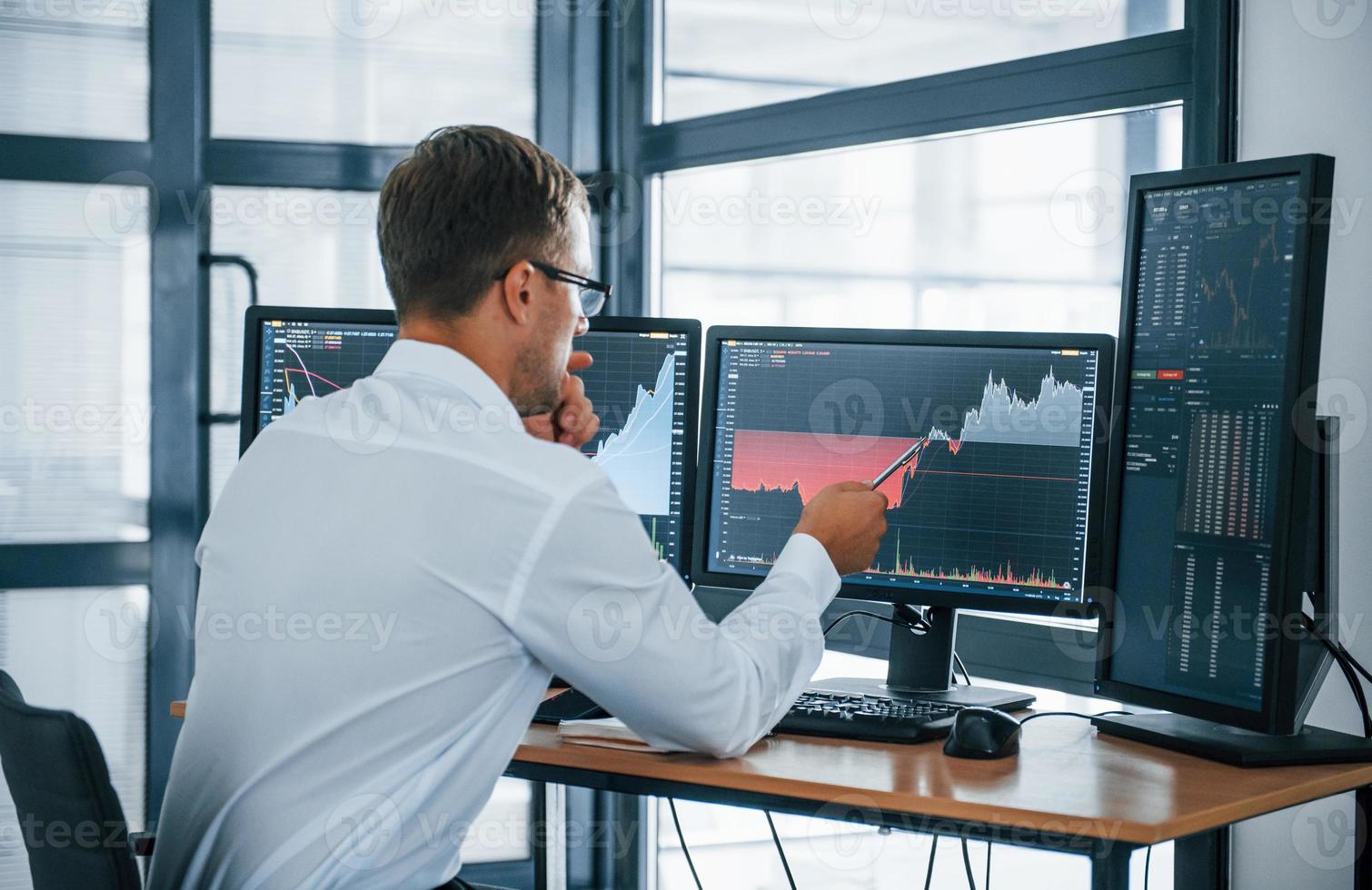 Looks at the graphs. Young businessman in formal clothes is in office with multiple screens. Conception of exchange and money photo
