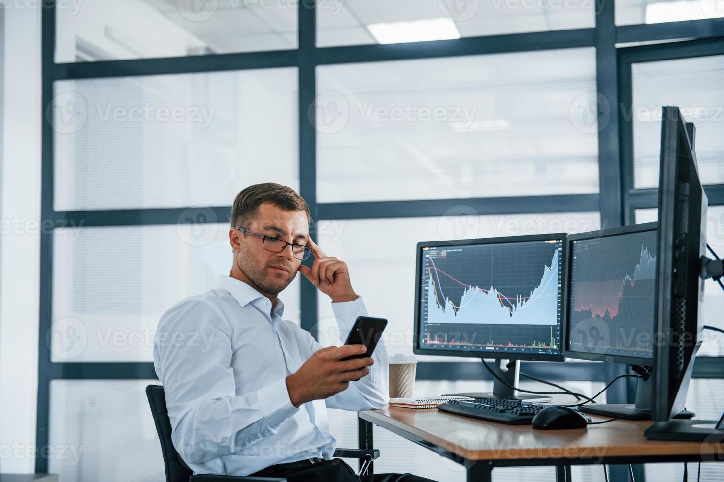 Using smartphone. Young businessman in formal clothes is in office with multiple screens. Conception of exchange and money photo