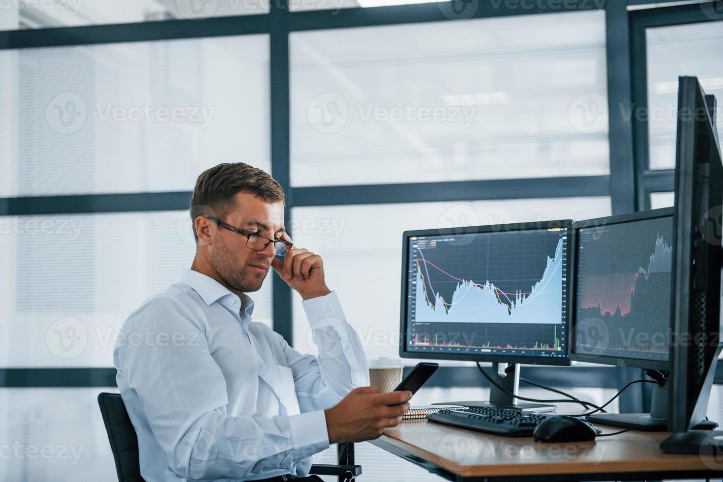 Using smartphone. Young businessman in formal clothes is in office with multiple screens. Conception of exchange and money photo