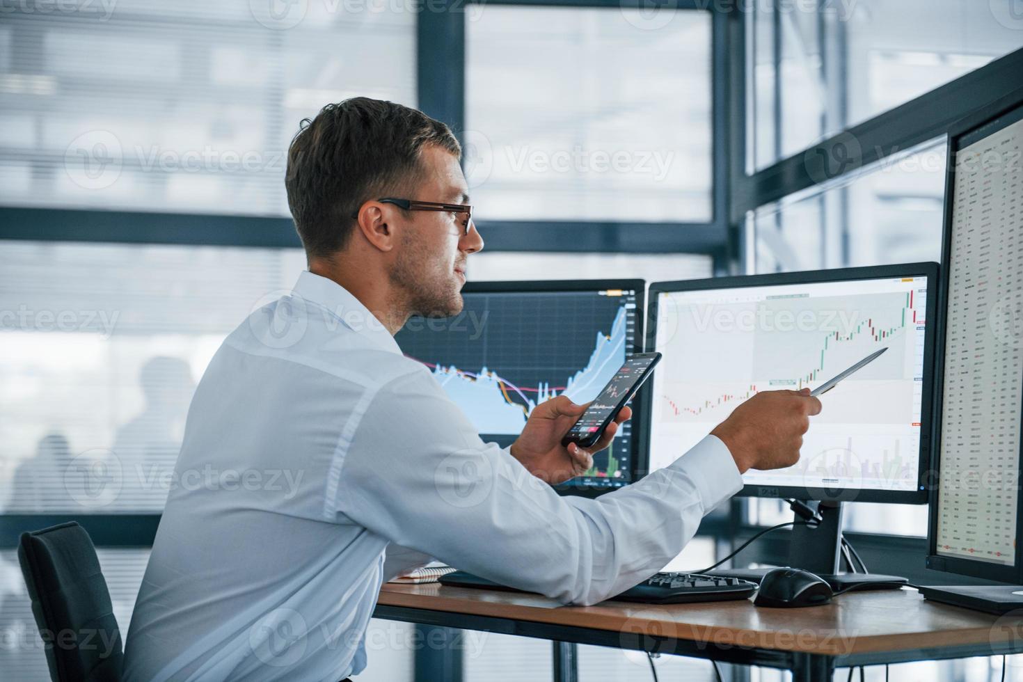 With phone and pen. Analysing graphs. Young businessman in formal clothes is in office with multiple screens. Conception of exchange and money photo
