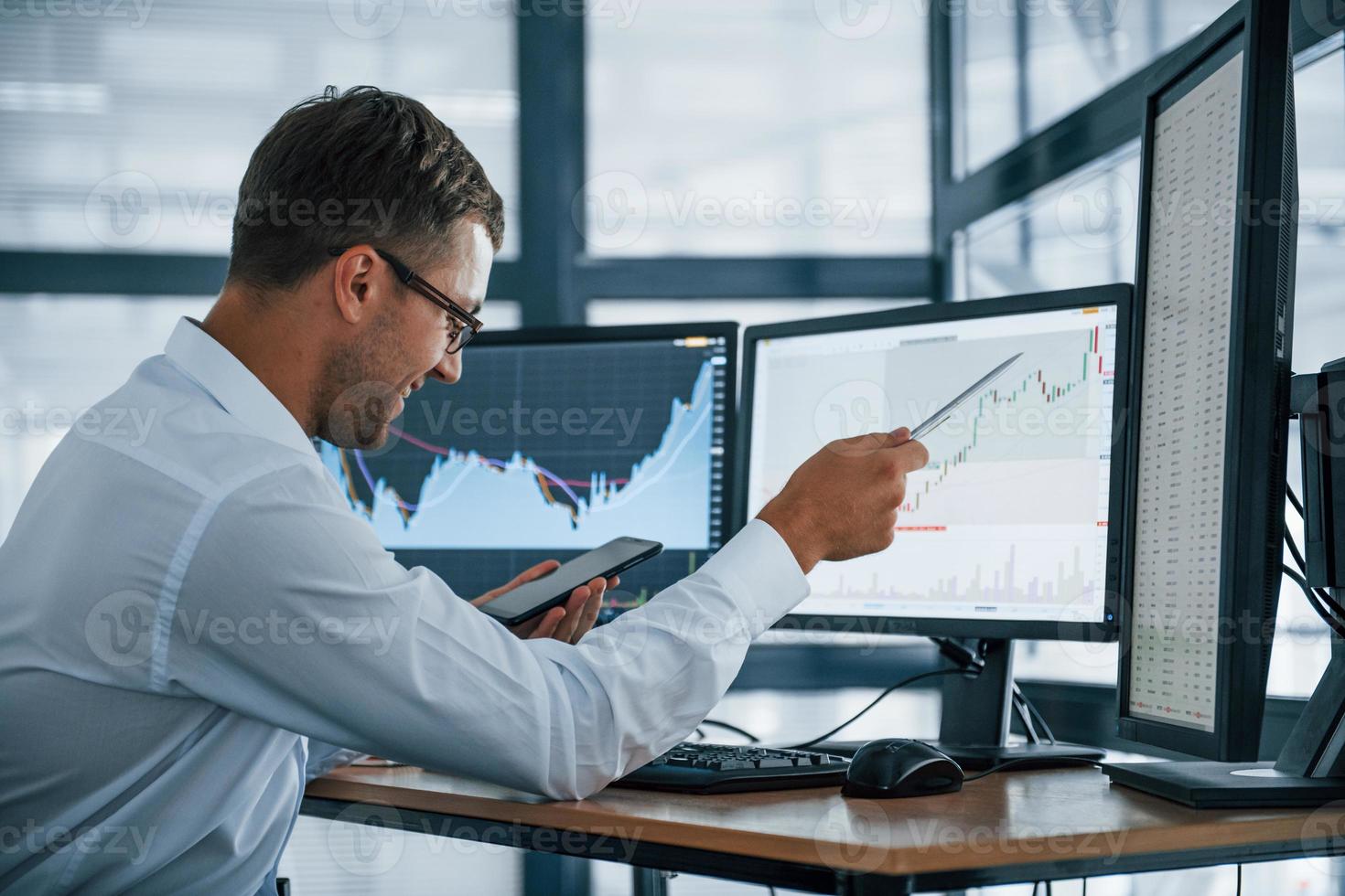 With phone and pen. Analysing graphs. Young businessman in formal clothes is in office with multiple screens. Conception of exchange and money photo