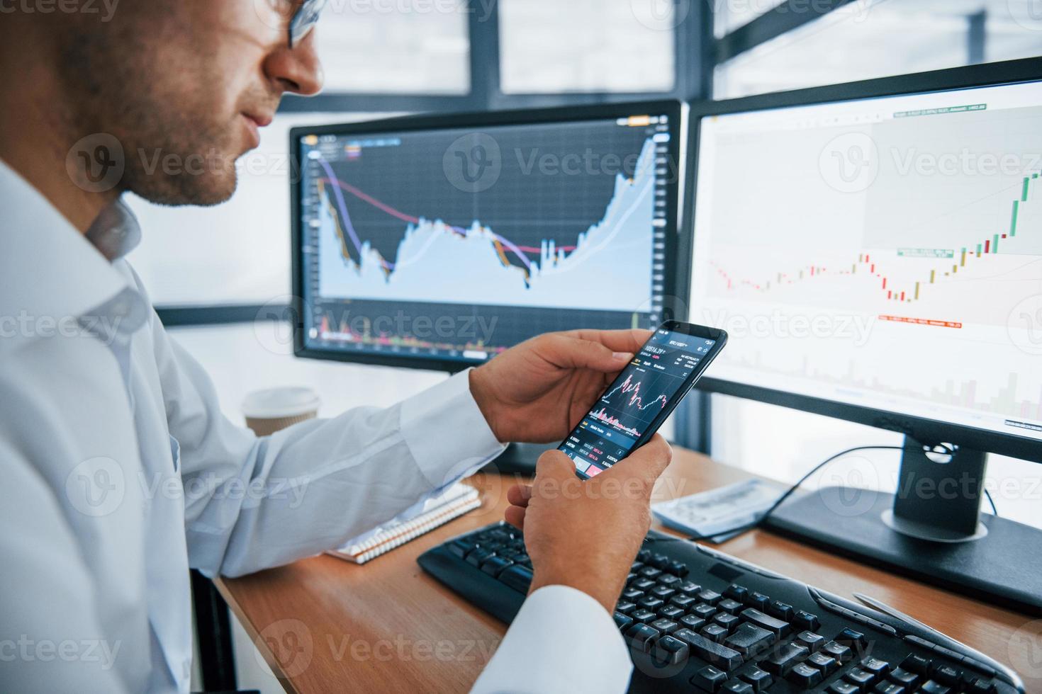 Using smartphone. Young businessman in formal clothes is in office with multiple screens. Conception of exchange and money photo