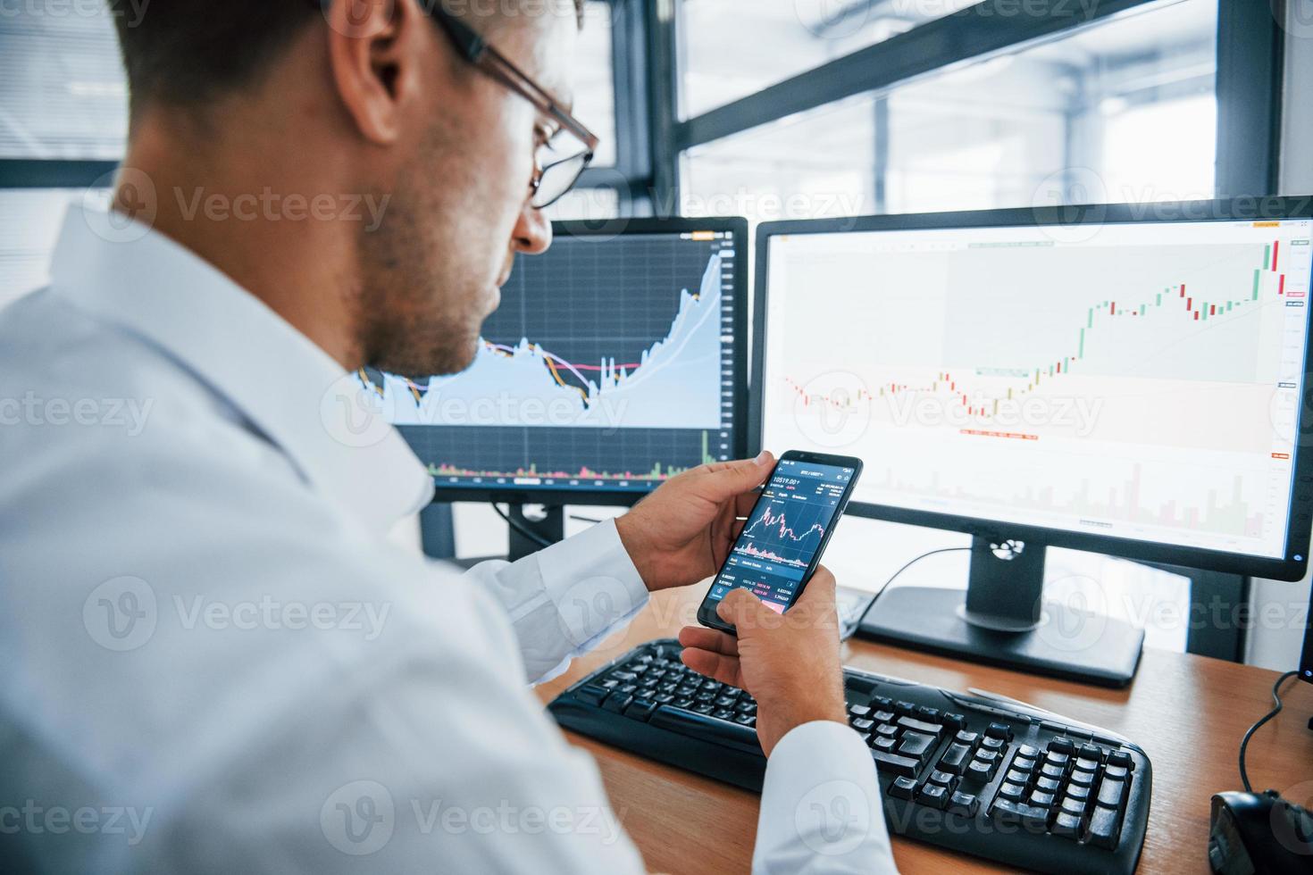 Using smartphone. Young businessman in formal clothes is in office with multiple screens. Conception of exchange and money photo