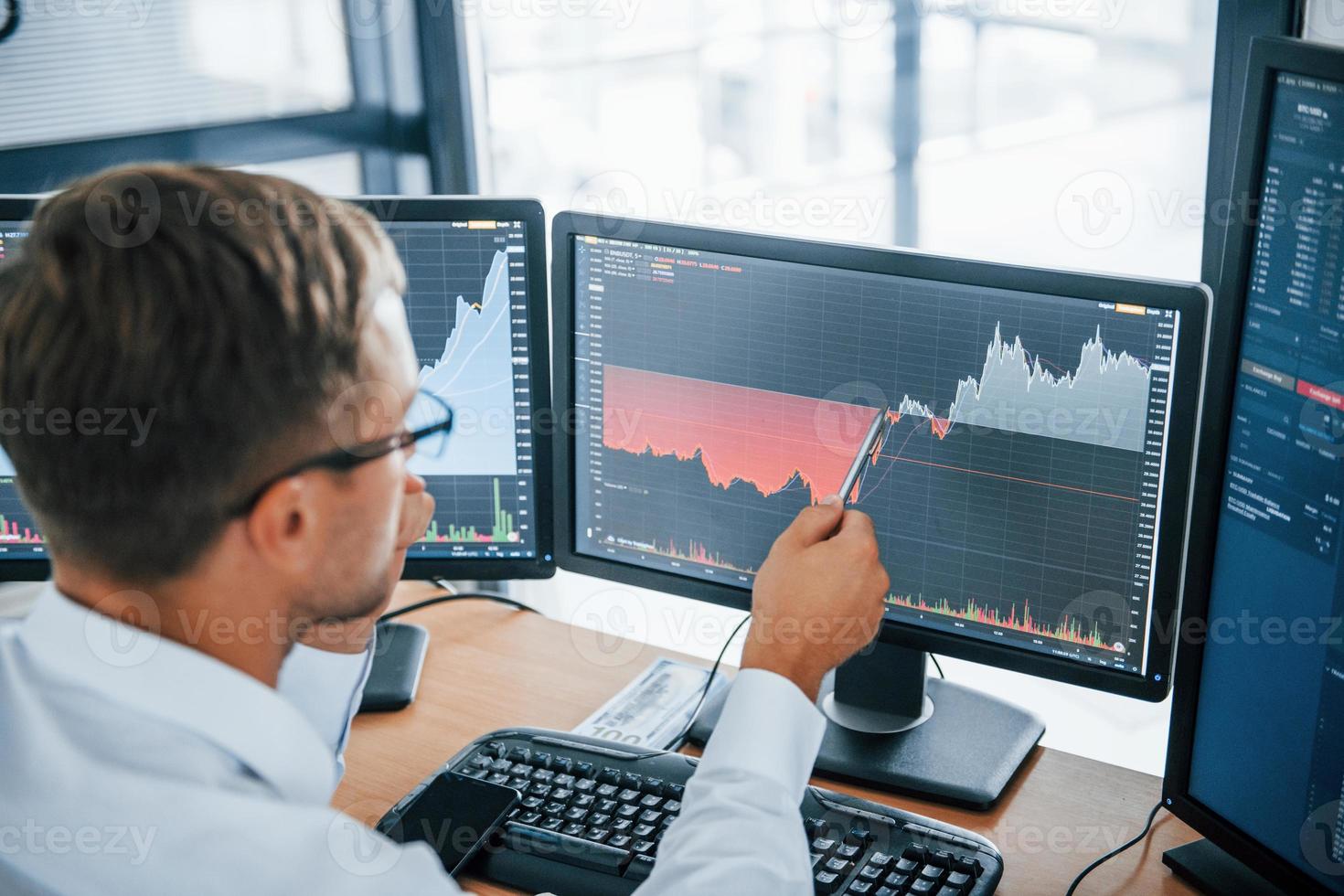 Looks at the graphs. Young businessman in formal clothes is in office with multiple screens. Conception of exchange and money photo