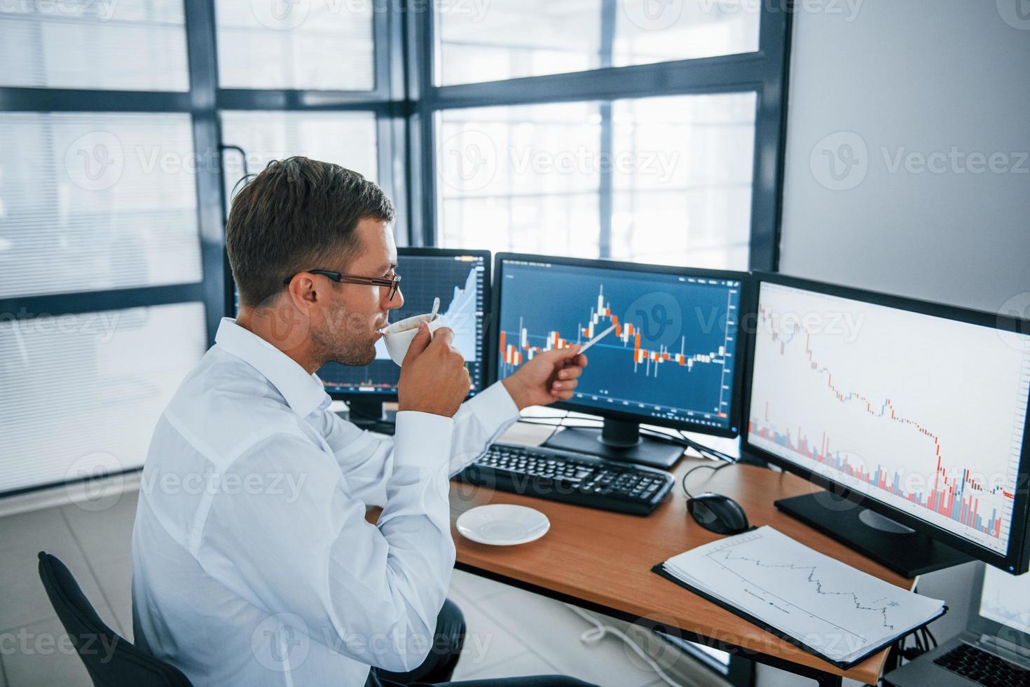 Holds cup of drink. Analysing information. Young businessman in formal clothes is in office with multiple screens. Conception of exchange and money photo