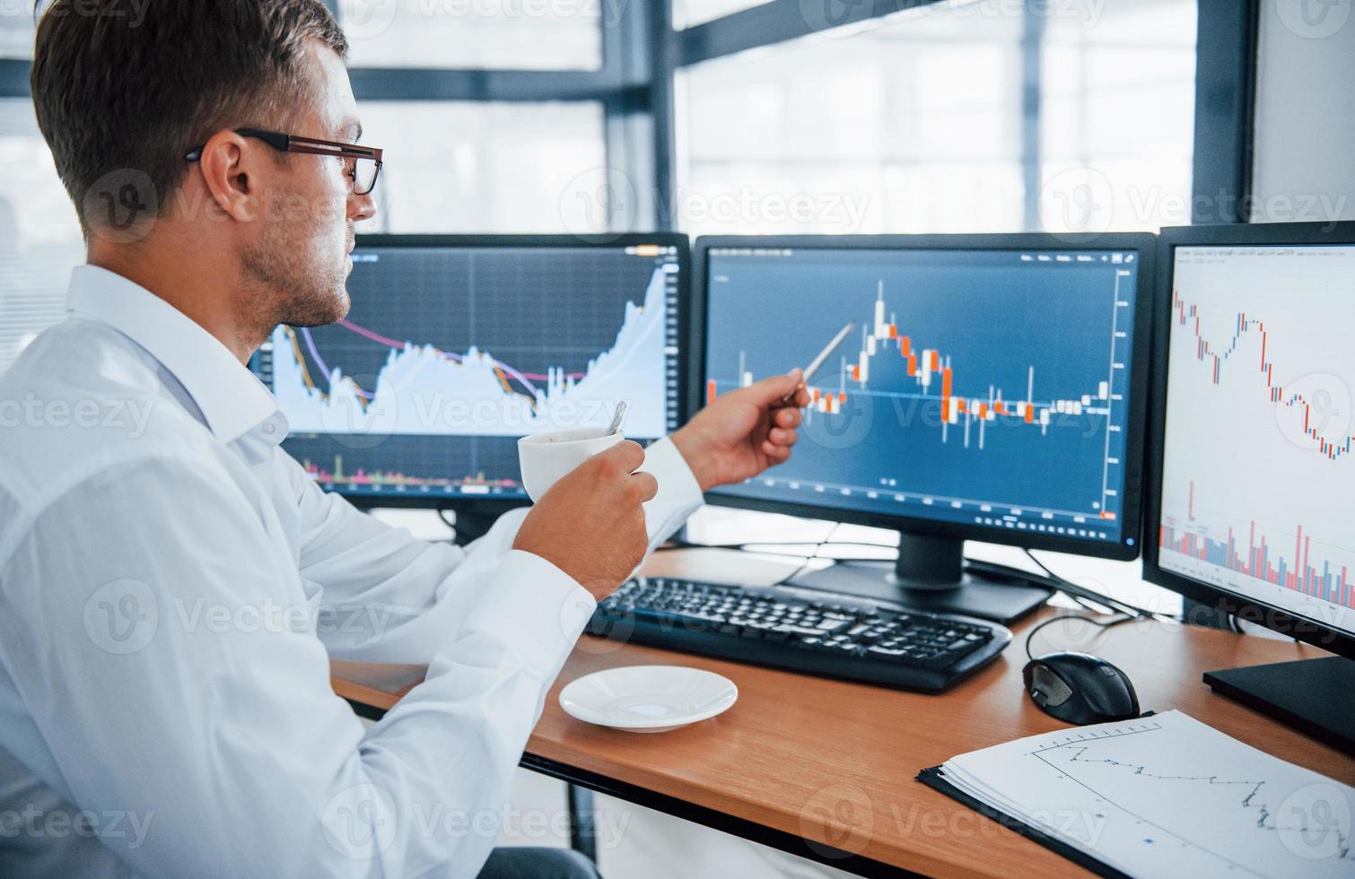 Holds cup of drink. Analysing information. Young businessman in formal clothes is in office with multiple screens. Conception of exchange and money photo