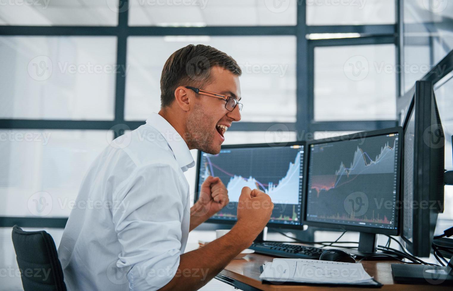 Excited and happy. Conception of victory and success. Young businessman in formal clothes is in office with multiple screens photo