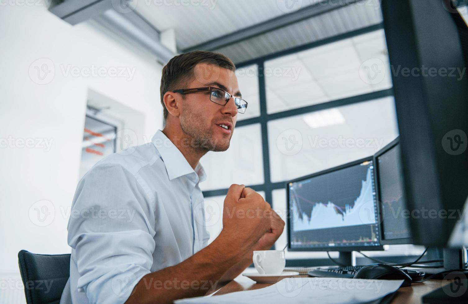 Excited and happy. Conception of victory and success. Young businessman in formal clothes is in office with multiple screens photo