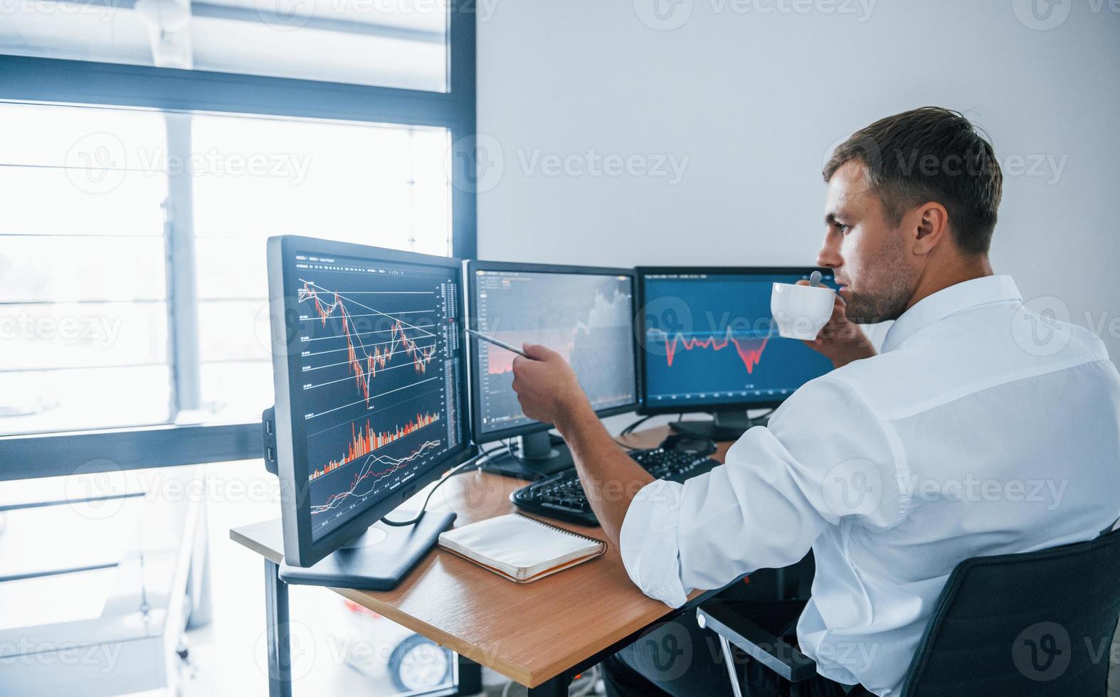 Young businessman in formal clothes is in office with multiple screens. Conception of exchange and money photo
