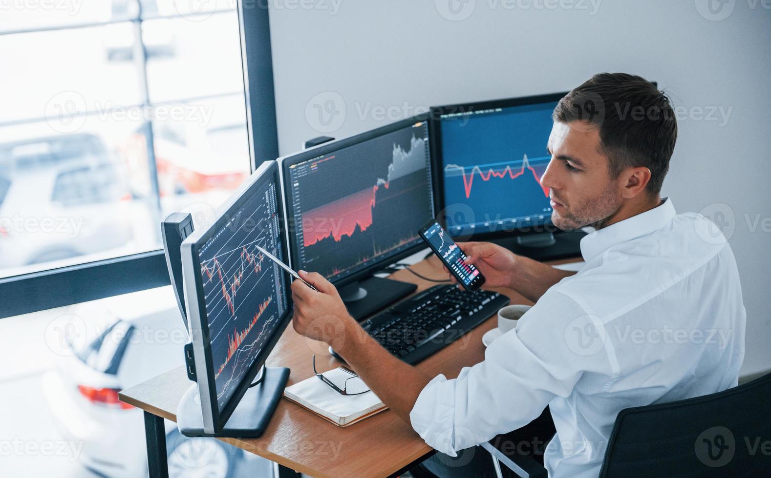 Young businessman in formal clothes is in office with multiple screens. Conception of exchange and money photo