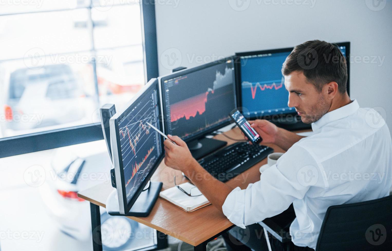 Young businessman in formal clothes is in office with multiple screens. Conception of exchange and money photo