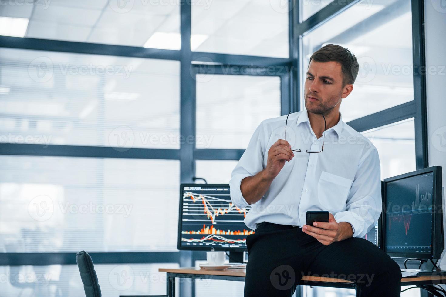 Sits on the table and having a break. With smartphone in hands. Young businessman in formal clothes is in office with multiple screens photo