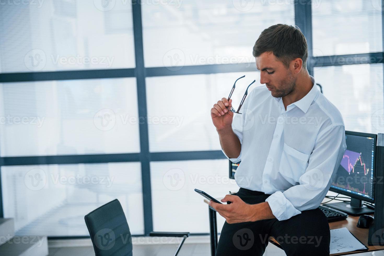 Sits on the table and having a break. With smartphone in hands. Young businessman in formal clothes is in office with multiple screens photo