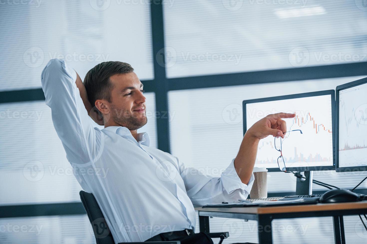 Feeling good. Conception of success. Young businessman in formal clothes is in office with multiple screens. Conception of exchange and money photo
